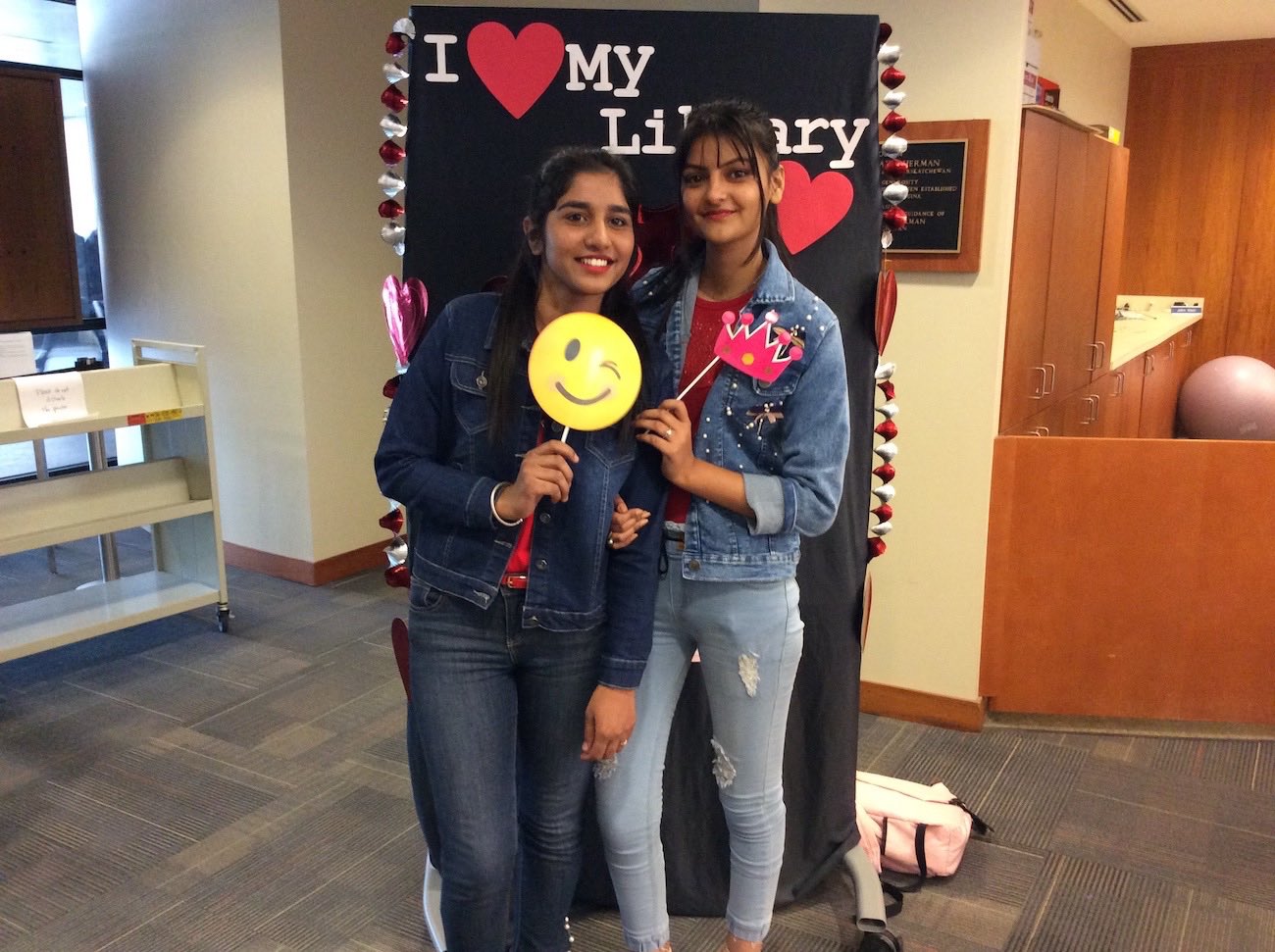 Two students with long black hair, wearing jeans and jean jackets, pose in front of the I love my Library backdrop each holding up a prop on a stick. The student on the left is holding a winking smiley face and the one on the right is holding a pink crown.